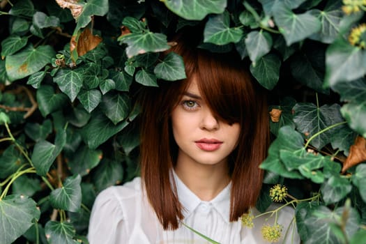 Redhaired woman in white shirt surrounded by lush green ivy foliage in outdoor setting