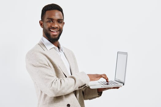 Cheerful man with dark skin, dressed in smart casual beige cardigan and white shirt, holding a silver laptop in hand, smiling confidently against plain white background, professional and friendly appearance. Concept: modern business success