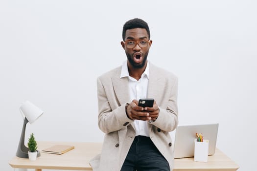 Happy man holding smartphone in bright modern office, surprised and excited mood, dressed casually in beige blazer and white shirt, office desk with laptop, stationery, minimal decor, clean white wall, workplace environment, lifestyle concept.