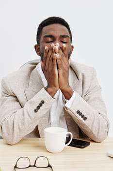 Tired and stressed young man with dark skin, closed eyes, holding nose, sitting at desk with coffee mug, smartphone, and glasses, representing fatigue, exhaustion, burnout, daily routine, indoor environment, neutral tone, casual clothing, emotional expression.