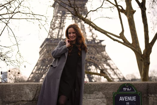 Editorial image of a woman in a grey coat standing near the Eiffel Tower on Avenue de New York in Paris, with winter trees and the Seine river in the background