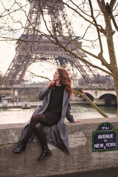 Editorial image of a woman sitting on a stone wall along Avenue de New York in Paris, smiling with the Eiffel Tower and the Seine river in the background on a cloudy day