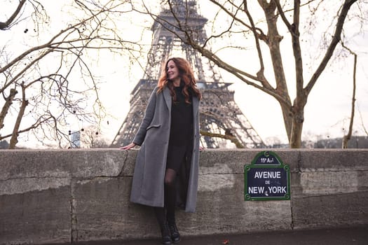 Stylish woman in a grey coat standing by the Seine river with the Eiffel Tower behind her on a cloudy day in Paris