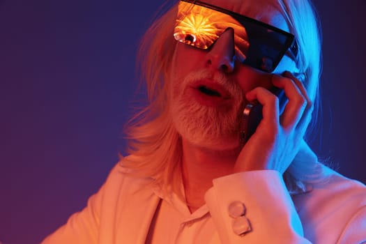 Cool man with long white hair dressed in a sharp white suit, wearing vibrant sunglasses that reflect a colorful sunset, speaking on his phone, embodies contemporary fashion and lifestyle