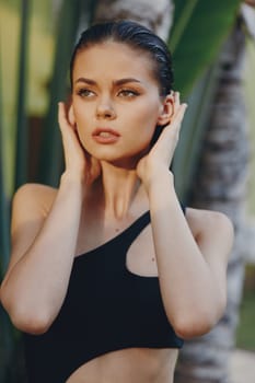Attractive young woman in fashionable black swimsuit posing in front of tropical palm trees for a photo shoot