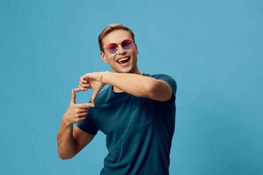 Smiling young caucasian man wearing blue t-shirt and pink sunglasses making frame gesture with hands isolated on plain blue background. Man expressing creativity and content generation using digital media. Concept: digital content creation.
