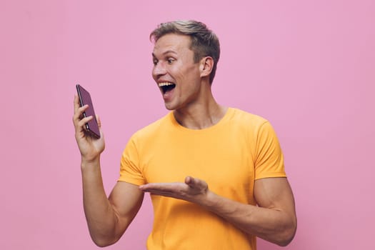 Cheerful man with short hair in yellow T-shirt looking at smartphone with happy expression isolated on pink background, showing surprise or excitement. Concept: social media, communication, technology