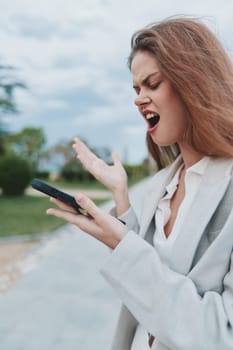 frustrated woman in casual business attire expressing anger and stress outdoors while looking at smartphone, symbolizing financial crisis and emotional struggle.