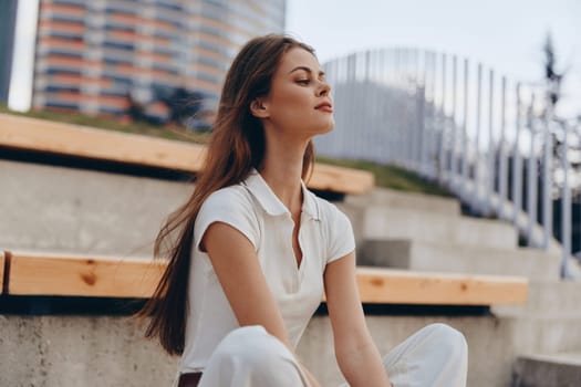 young woman in casual white clothing with long hair sits on urban steps enjoying a calm and peaceful moment in natural daylight, capturing a serene outdoor lifestyle scene