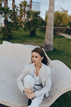 Young woman in light suit sits thoughtfully on curved stone bench outdoors in spring park, evoking fashion and psychological concept with natural green background.