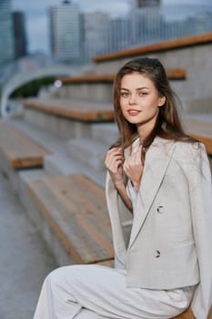 Stylish young woman wearing light blazer and white pants sitting on urban stairs, showing confidence and elegance with natural makeup and soft daylight, fashion style
