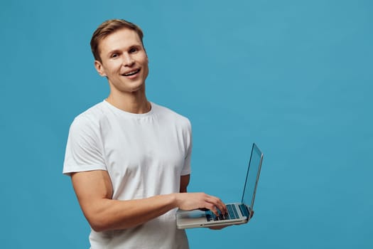 Happy young man in casual clothing holding a laptop computer against a plain blue background, smiling at camera, relaxed pose, technology and communication concept, studio portrait, modern lifestyle, digital device, cheerful expression, casual fashion.