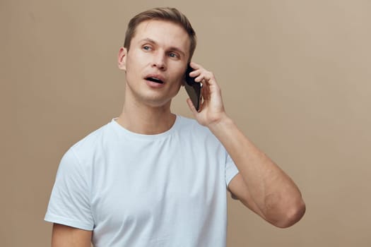 Close-up of a smiling young man with short hair, casually dressed in a light blue t-shirt, holding a mobile phone to his ear, reacting with astonishment as he talks, against a neutral beige backdrop. Concept: communication and surprise
