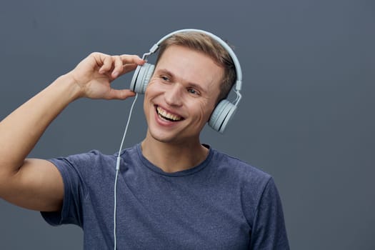 Happy young man enjoying music through headphones, casual blue t-shirt, positive mood, gray studio background, close-up portrait, leisure, entertainment, relaxation concept.