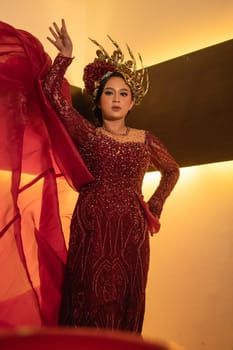 A glamor woman poses with a flying red dress on her body while wearing a gold crown inside the cream studio