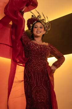 A glamor woman poses with a flying red dress on her body while wearing a gold crown inside the cream studio