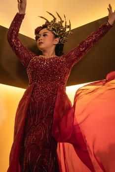 A glamor woman poses with a flying red dress on her body while wearing a gold crown inside the cream studio