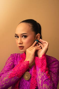Asian woman in a pink dress trying to wear a green earring in her ears inside the studio