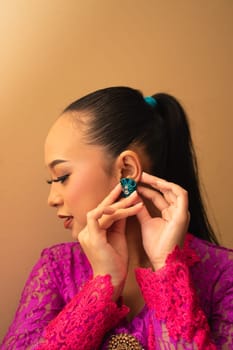 Asian woman in a pink dress trying to wear a green earring in her ears inside the studio
