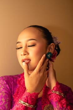 Asian woman in a pink dress trying to wear a green earring in her ears inside the studio