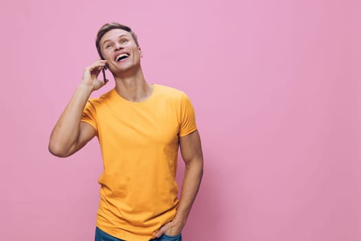 Smiling young man talking on phone wearing orange t-shirt standing against pink background expressing cheerful positive emotion. Casual lifestyle communication digital nomad concept with mobile phone technology.