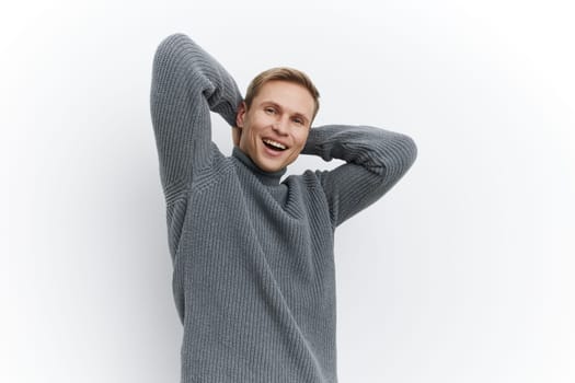 Cheerful young man with blond hair dressed in a warm gray knitted sweater smiles joyfully at the camera, expressing happiness and confidence. Isolated on a plain white background, this portrait highlights positive mood and casual style. Concept: joy of everyday life