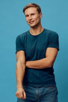 Happy young man with short hair in casual teal t-shirt posing with crossed arms and smiling at the camera on a solid blue background, studio setting, lifestyle image capturing positivity and confidence