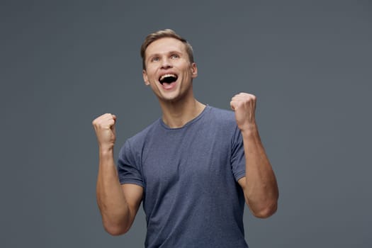 Happy man feeling victorious and energized, casual outfit, smiling with enthusiasm, celebrating achievement, plain gray backdrop, studio environment, positive expression. Concept: success and happiness.