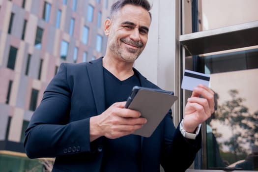 Confident caucasian man with headphones smiling while using tablet and holding a credit card outdoors. Concept of online shopping, digital payment, modern lifestyle, and financial technology.