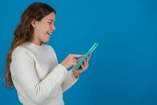 Profile of a smiling young woman touching the screen of a green digital tablet against a solid blue background with copy space, symbolizing interaction, technology, and modern communication gesture.