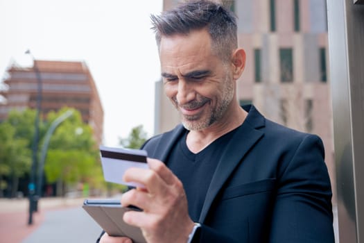 Confident caucasian man with headphones smiling while using tablet and holding a credit card outdoors. Concept of online shopping, digital payment, modern lifestyle, and financial technology.