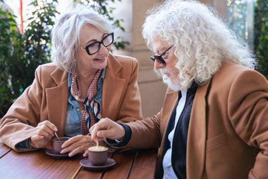 Caucasian senior couple enjoying coffee at an outdoor cafe. The woman speaks closely to the man, creating an intimate and thoughtful moment. Concept of connection, trust, and shared experiences.