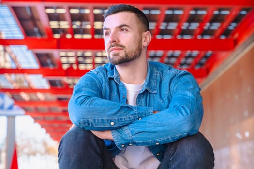 Portrait of a caucasian young man sitting thoughtfully in a modern city environment. Casual denim outfit, red architectural elements above, and natural light create a stylish and relaxed urban vibe.