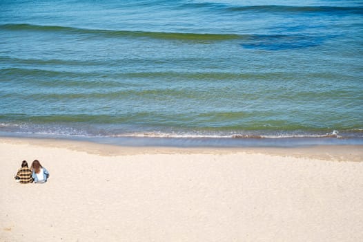 Two girls in clothes are sitting on a sandy beach and looking at the sea. Clear blue sky, calm sea. The girls are sitting with their backs to the camera. A group of people on a beach.