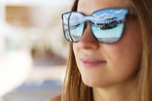 Close-up of woman wearing sunglasses showing reflection of a water park with pool and slides on a sunny day.