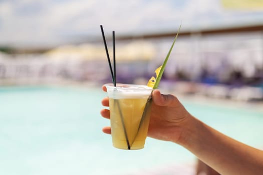 A hand holding a refreshing tropical cocktail in a plastic cup with straws and garnish, set against the backdrop of a sunny poolside. Perfect for summer vacation and leisure themes.