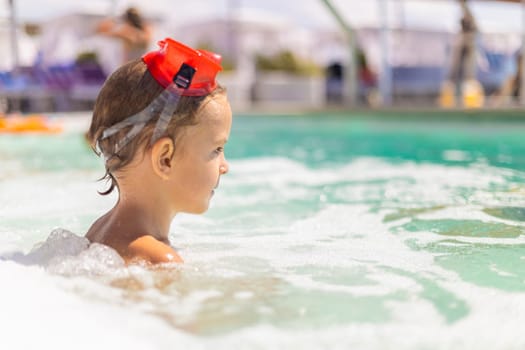 Young child with red swim goggles sitting in shallow foamy water at a pool with blurred lounge chairs in background.