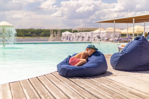 A young child wearing a cap and striped shorts is peacefully napping on a blue bean bag next to a swimming pool. The scene captures a tranquil summer day with clear skies and a serene poolside atmosphere.
