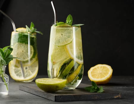 Refreshing detox or summer drink in a glass with lemon, lime, mint, ice, and metal straw. Dark background