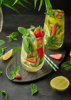 A refreshing drink in a glass with lemon slices, strawberries, fresh mint leaves, and sparkling water, served with lime and mint on a black plate