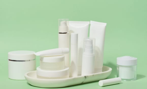 A collection of various white cosmetic containers, including tubes, jars, and bottles, are arranged on a white stand against a light green background 