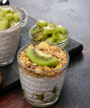 Two glasses of chia seed pudding, one topped with diced kiwi and the other with granola and banana chips, are on a dark surface with ingredients