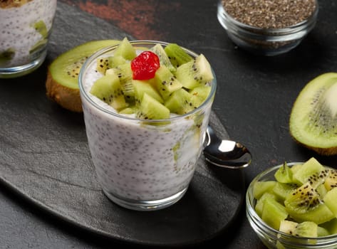 Glass of chia seed pudding topped with diced kiwi and a red cherry sits on a dark surface with halved kiwis, a bowl of diced kiwi, and chia seeds