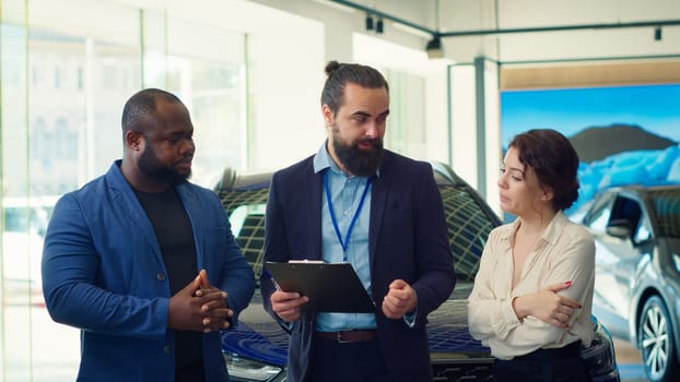 Dealership salesman answering customers questions about cars, financing and purchase process. Vehicle showroom agent explaining clients warranties, services and available extra features, camera A