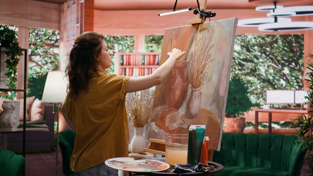 Woman artist wiping off a color from her paintbrush before mixing oil paints, creating multicolored rustic artwork with paint tubes. Painter showing talent and creativity in art studio. Camera B.