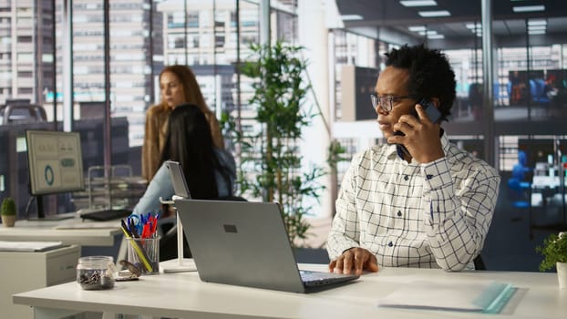 Businessman engages in a mobile phone conversation at his desk, managing a project and discussing deal making strategies. Multitasking skills drive productivity in a corporate setting. Camera B.