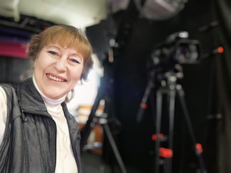 A middle aged female blogger smiles for a selfie while getting ready for a lifestyle video shoot in a lively studio environment.
