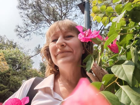 A middle-aged blonde woman smiles as she plays with vibrant pink flowers, capturing a joyful moment in a sunny garden.