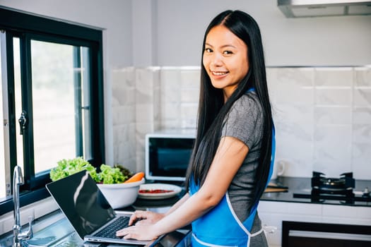 A woman stands in the kitchen happily cooking and using a laptop for tutorials or recipes. Illustrating the joy of technology in culinary exploration and meal preparation.
