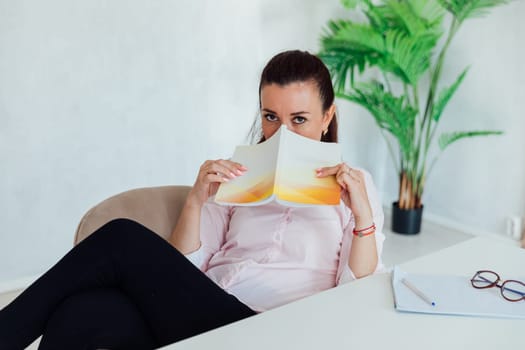 brunette woman at office desk covering her face with a book